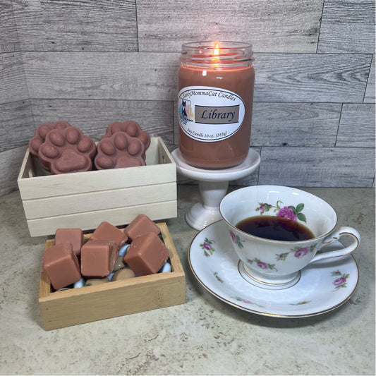 Brown Library soy glass jar candle lit on a white pedestal, wax melt cubes in a little wooden box, paw print wax melts in a wooden box and a flowery patterned tea cup and saucer. .