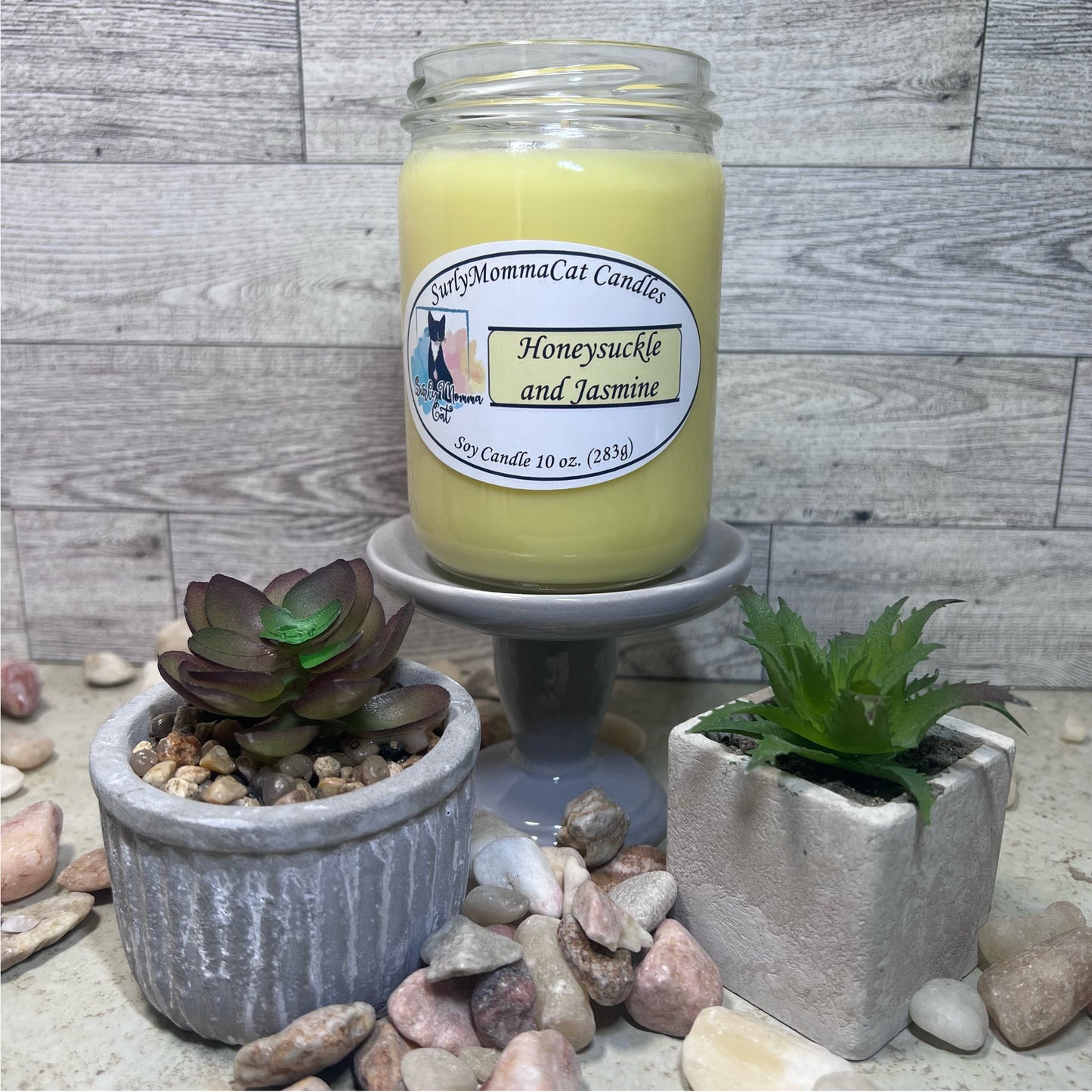 Yellow Honeysuckle and Jasmine soy glass jar candle on a gray pedestal with rocks and plants for decoration.