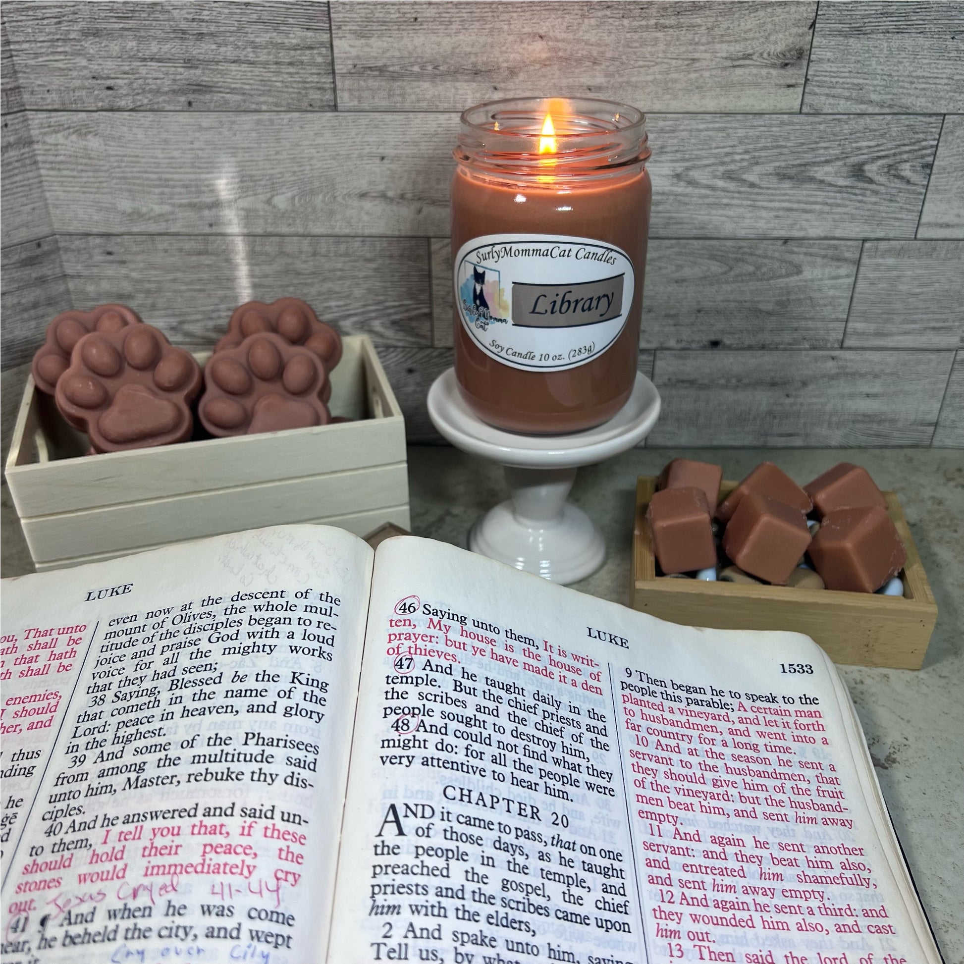 Brown Library soy glass jar candle lit on a white pedestal, wax melt cubes in a little wooden box, paw print wax melts in a wooden box and the Holy Bible open to the book of Luke. 