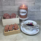 Brown Library soy glass jar candle lit on a white pedestal, wax melt cubes in a little wooden box, paw print wax melts in a wooden box and a flowery patterned tea cup and saucer. .
