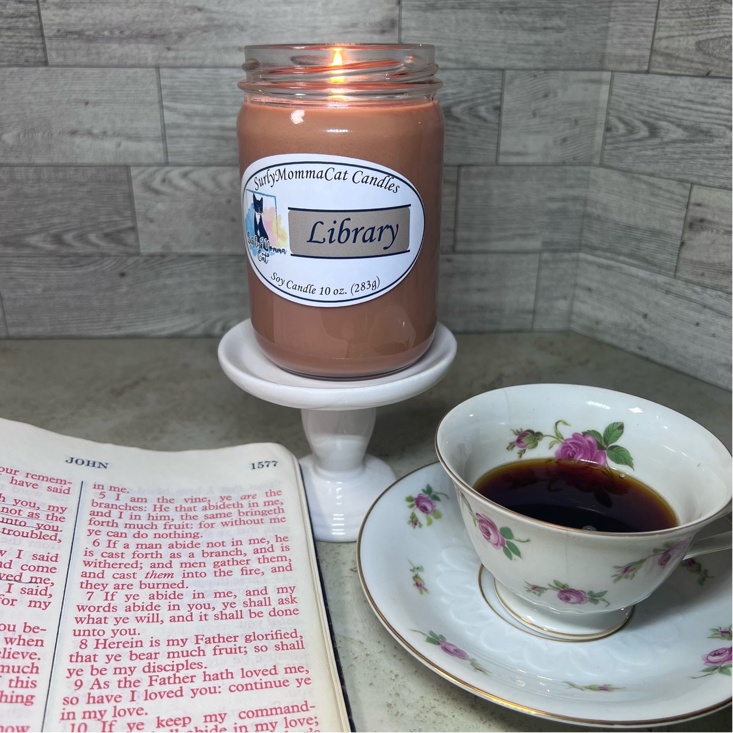 Brown Library soy glass jar candle lit on a white pedestal, and the Holy Bible open to the book of John, with a flowery patterned tea cup and saucer..