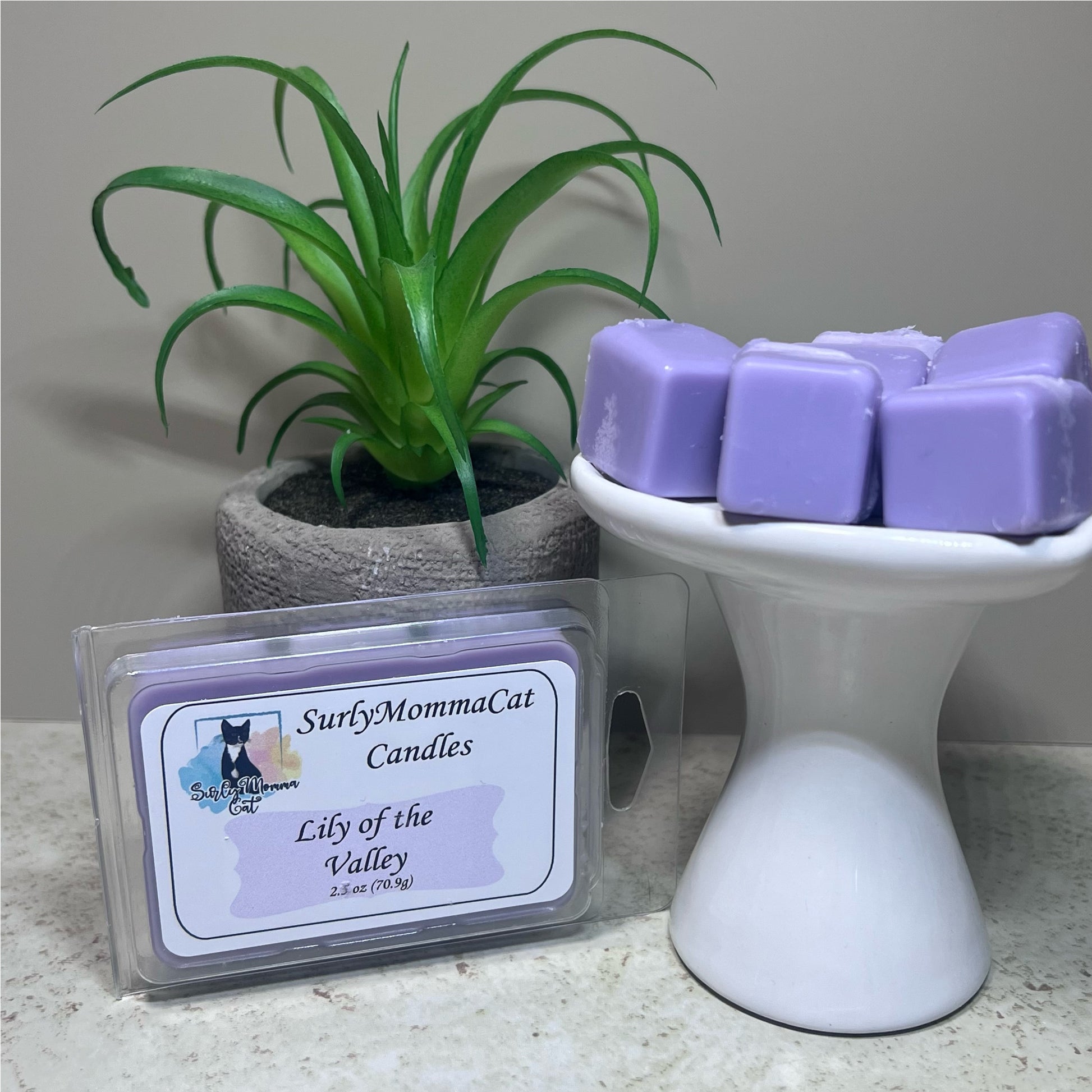 Light Purple Lily of the Valley Soy Wax Melts on a small white pedestal, and in a box labeled Lily of the Valley, with small plant for décor.
