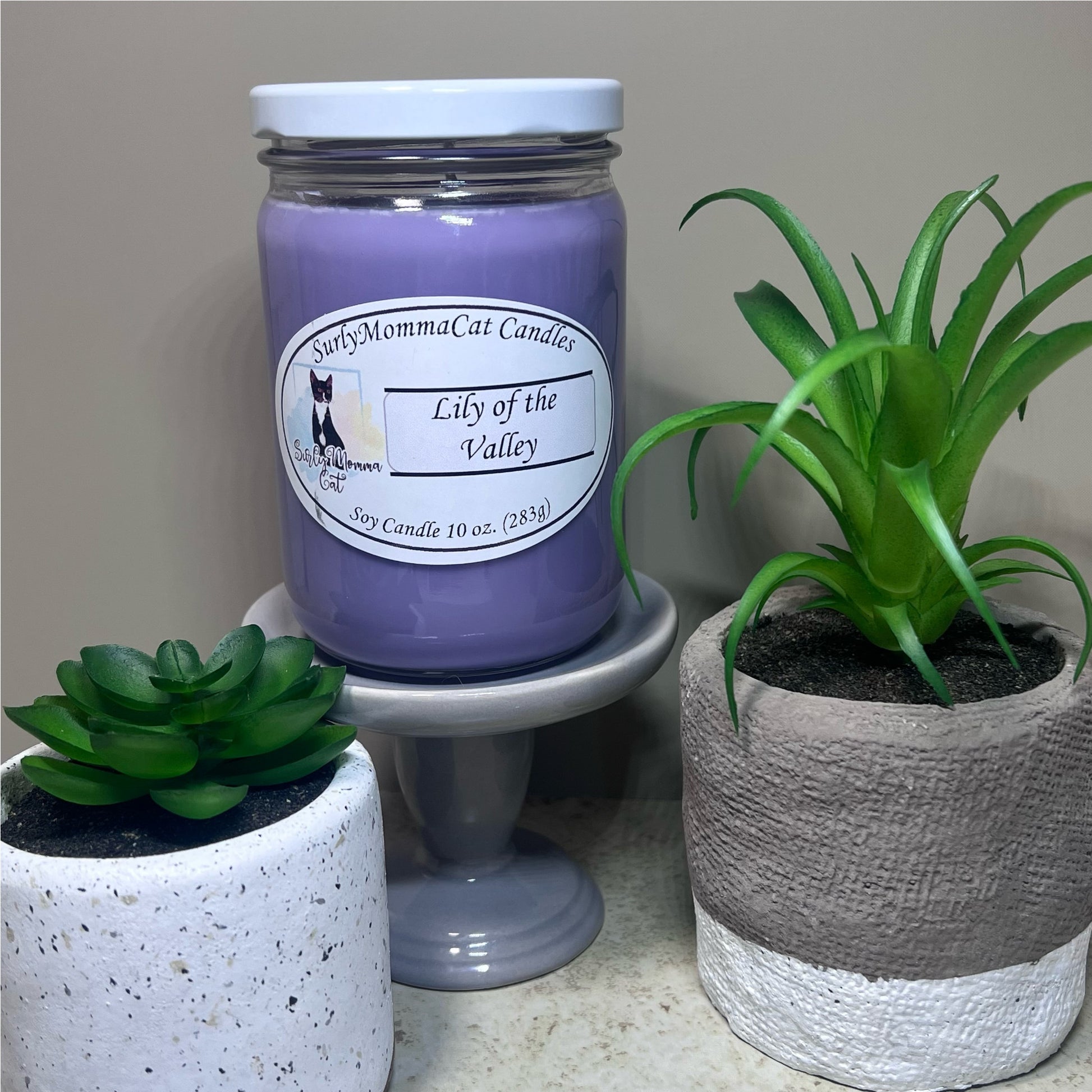 Light Purple Lily of the Valley Soy Glass Jar candle, on small white pedestal, with a small plants for décor.