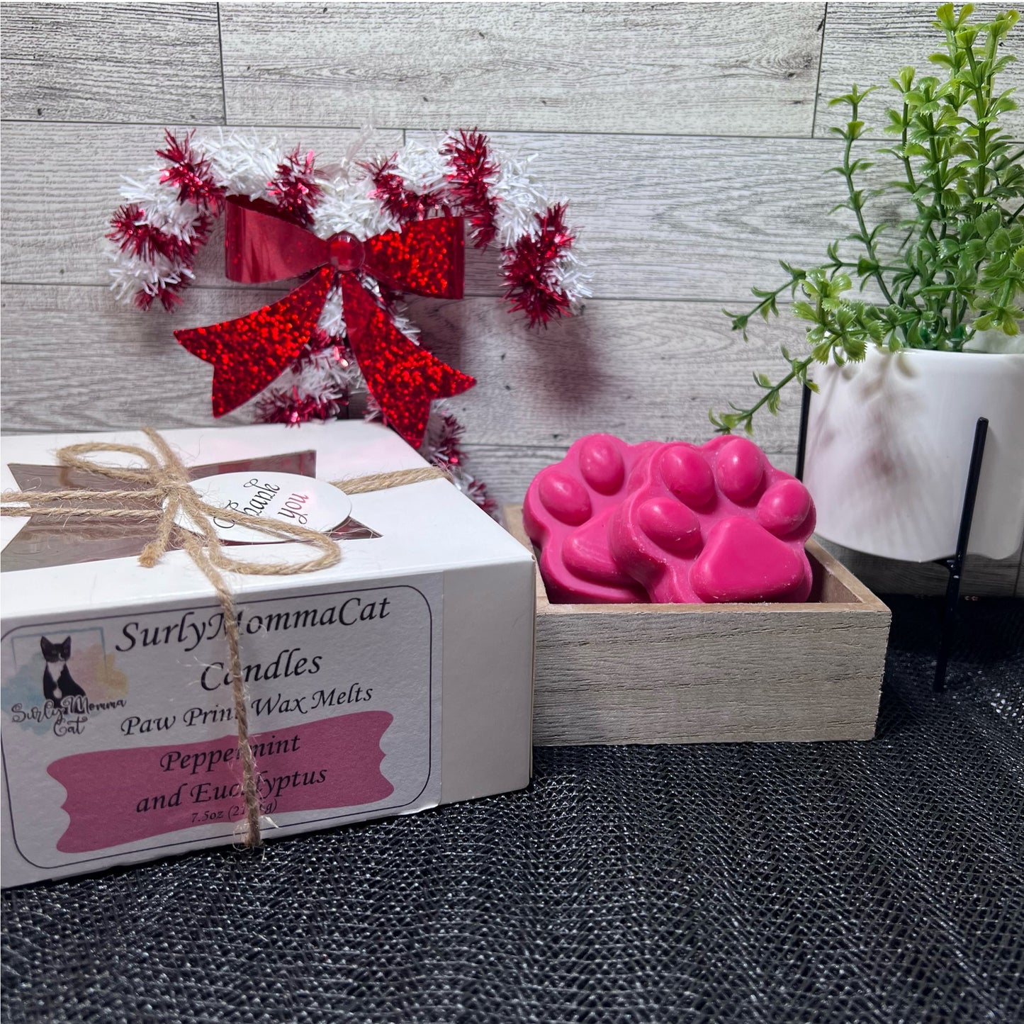 A warm scented Peppermint and Eucalyptus Christmas scene, with the featured wax melt candles in the adorable paw print design, accenting the scene as background decoration stands a lovely green plant budding with flowers, and a red and white Christmas candy cane wreath.