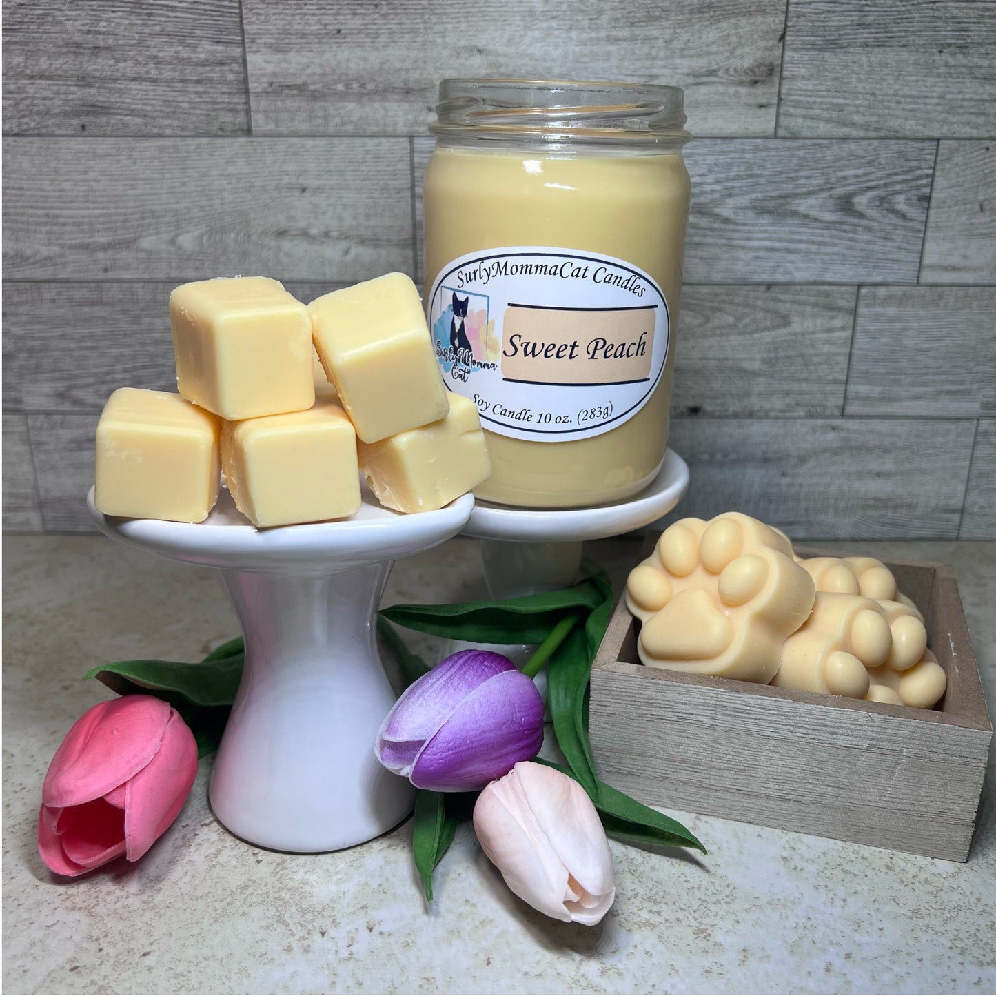 Tan Sweet Peach soy glass jar candle with cubed and pawprint wax melts on small white pedestal with the paw prints in a rustic box, with some pink, purple, tan flowers for decoration.